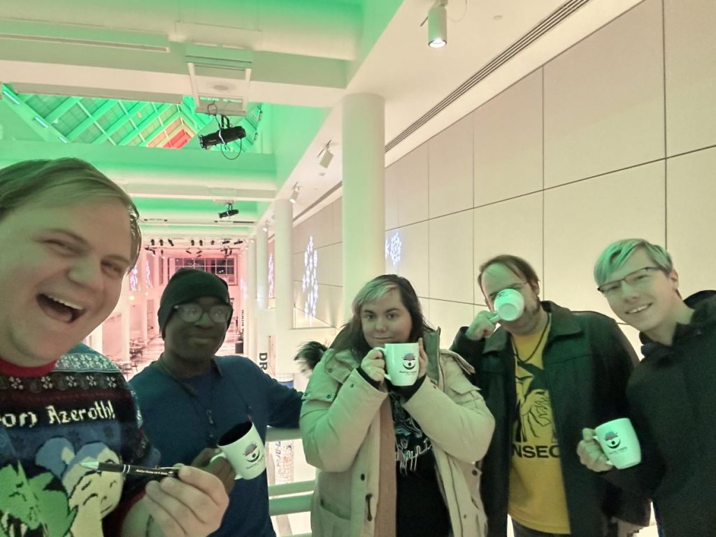 An image of 5 Amatryx lab members with coffee mugs and pens with the Amatryx logo on them. The group is in the atrium of the Center for the Arts at UB, with holiday colored lights in the background.