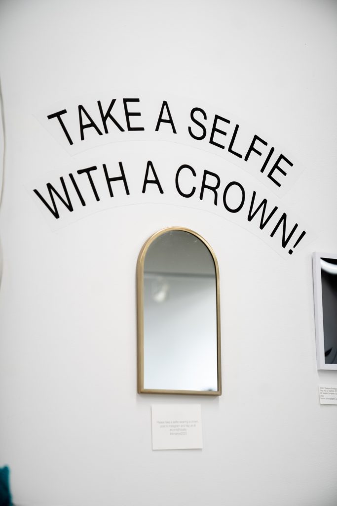 An image of the mirror on the wall of the exhibition, with text instructions to take a selfie with a crochet crown. 