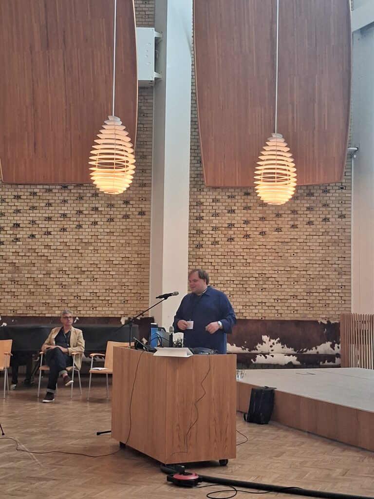 An image of Cody Mejeur delivering their talk as a Contemporary Narrative speaker at the Narrative 2026 conference in Aarhus, Denmark. Cody is wearing a blue robe and standing at a desk and microphone at the front of a large hall.