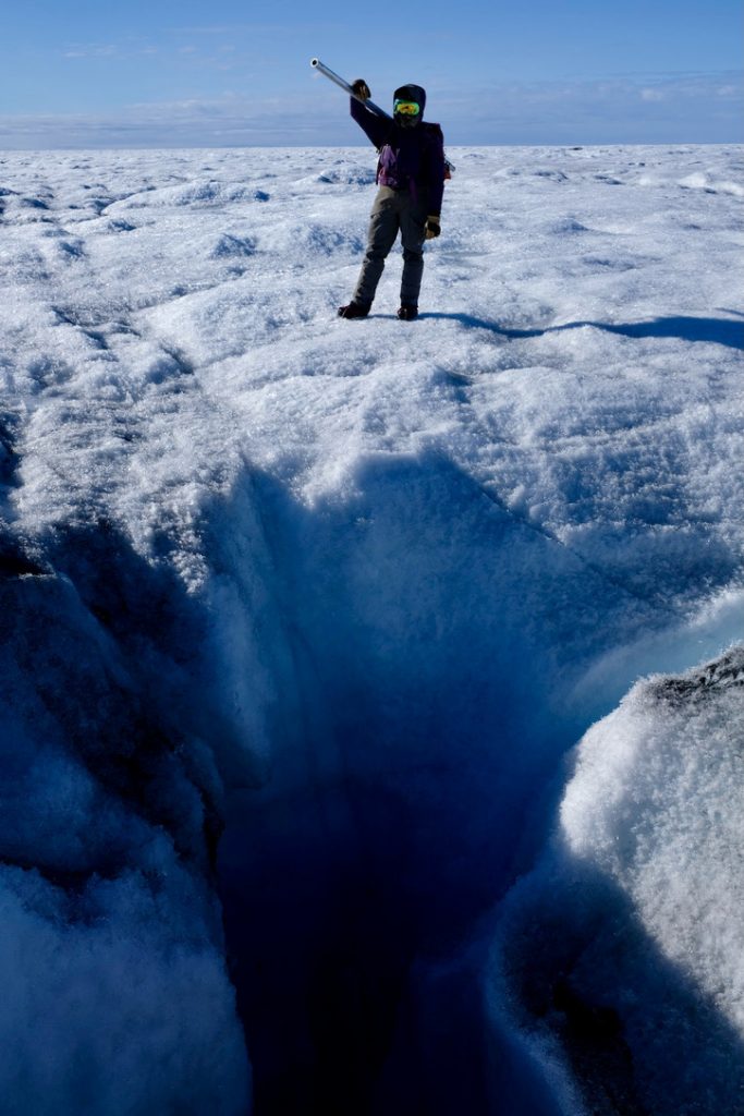 Person standing on bare ice with a moulin in the foreground.