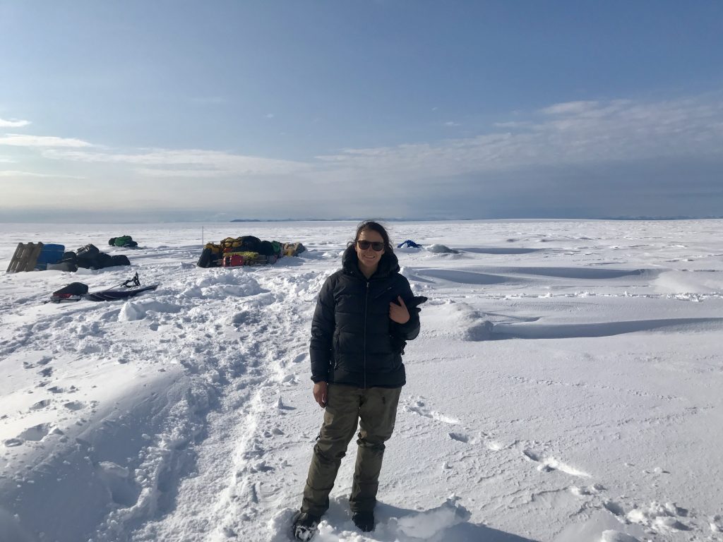 woman in a navy parka and tan snow pants standing on a a flat snow-covered surface (Greenland Ice Sheet) Blue sky with clouds in the distance