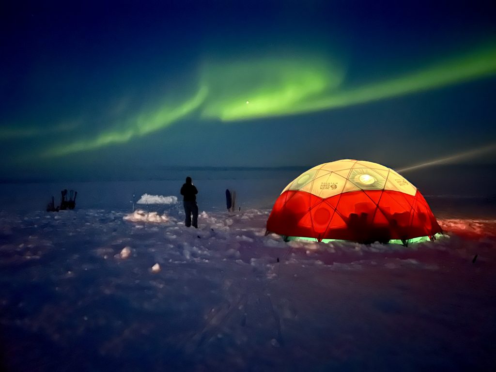 An illuminated domed orange and white tent is pictured at night with the northern lights in green lighting up the sky, snow covers the ground and two people can be seen in the distance