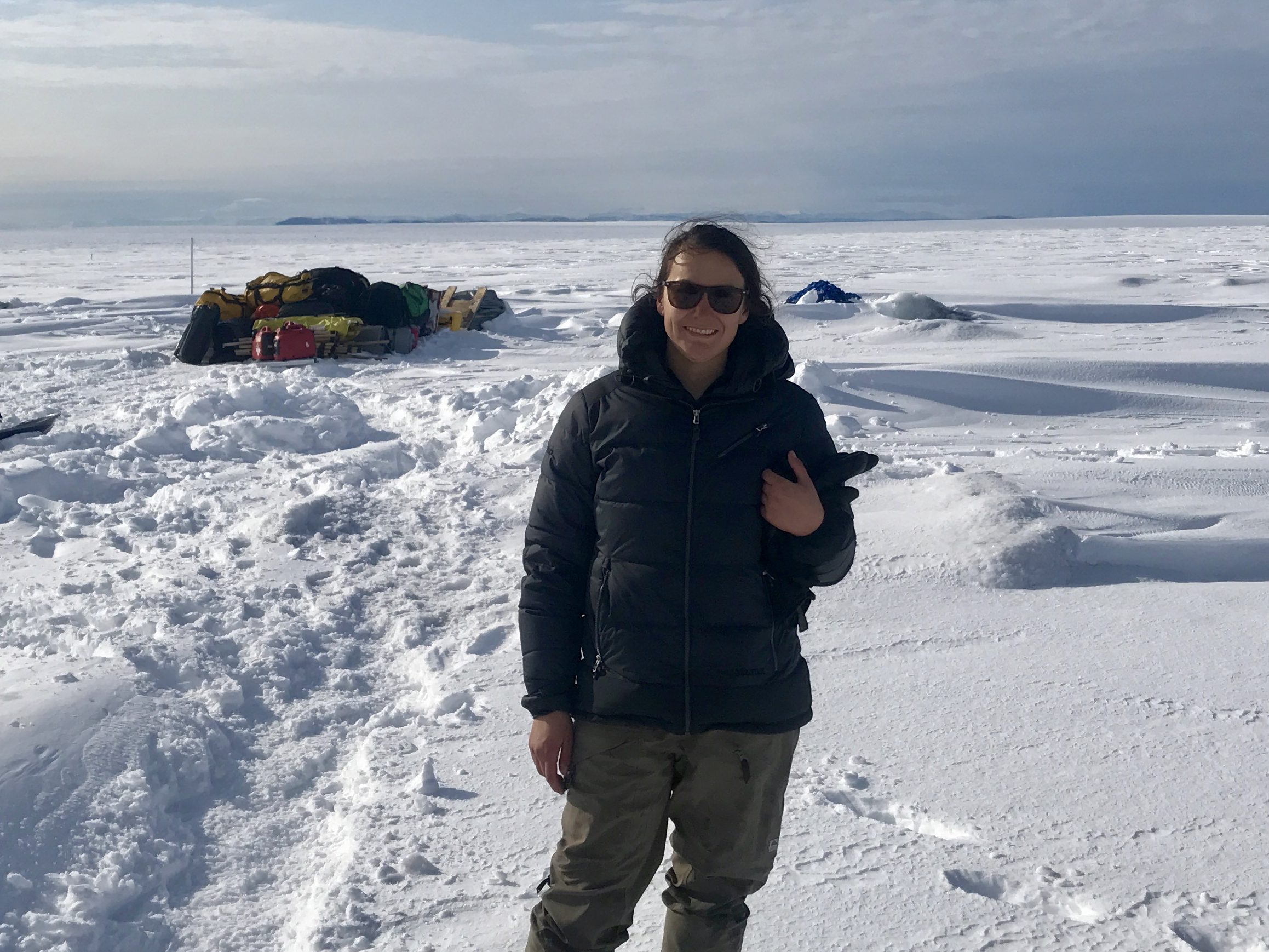 woman in a navy parka and tan snow pants standing on a a flat snow-covered surface (Greenland Ice Sheet) Blue sky with clouds in the distance