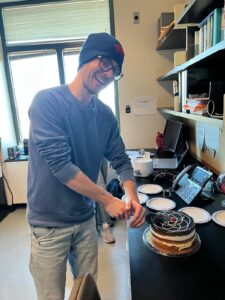 A grinning Professor Lacy, wearing UB blue and Buffalo Bills beanie, slices a cake. In the background of his mind, however, is the pestering question about how to make MnCl3