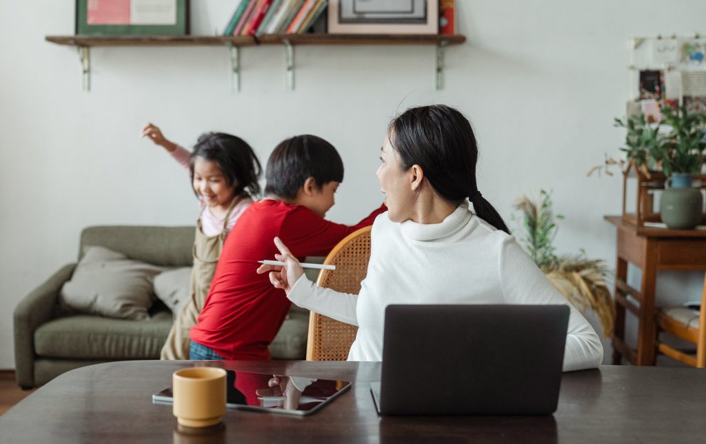 Mom works on laptop while kids play behind her.