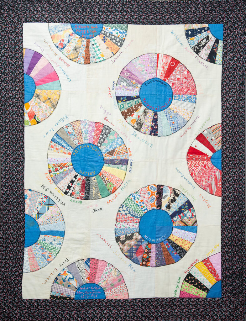 The Jensen family quilt’s twelve squares hold blue plates within larger circles of bright calico fabrics. Handstitched names of family members surround each circle and are slightly bumpy to the touch. The quilt is wide and long enough to wrap around two people. This piecework was made by Prairie Band Potawatomi healer O-Zoush-Quah, and her daughter, Pah-Kish-Ko-Quah, while O-Zoush-Quah was incarcerated at Canton Asylum, ca. 1910–30.