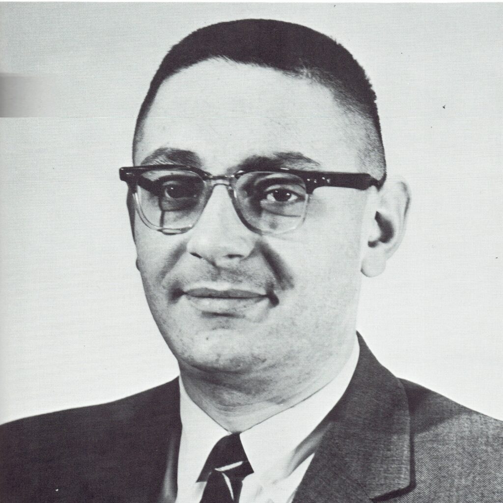 A picture of Dr. Milton Plesur. The photo is black and white, and the man's head and shoulders are visible. A white man with dark hair shaved close to the head looks at the camera, slight, closed-mouth smile on his face. He is wearing rounded, wire-framed glasses. He is wearing a dark suit jacket, white collared shirt, and a dark tie with white diamond stripes partially visible in frame.